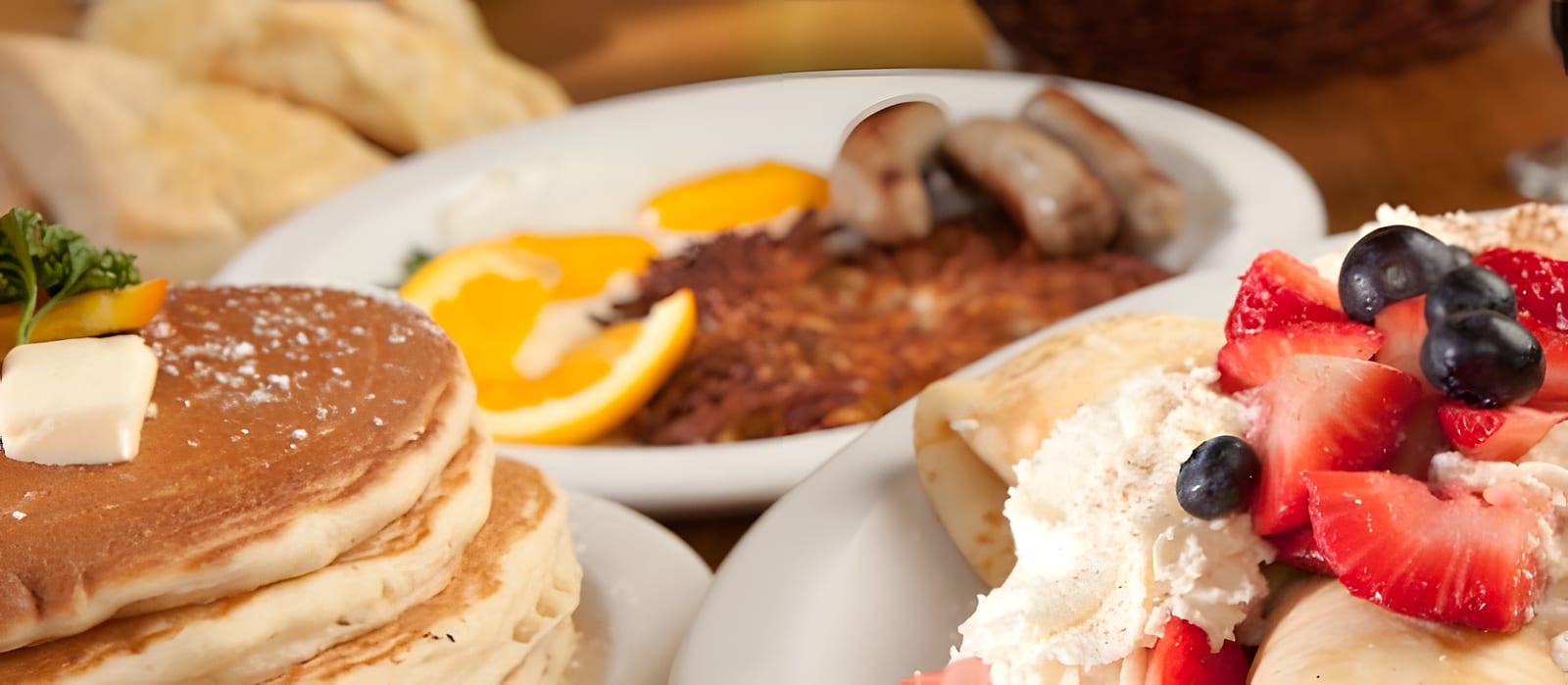 A stack of pancakes with butter beside a berry-topped crepe and a plate of eggs, sausage and hash browns