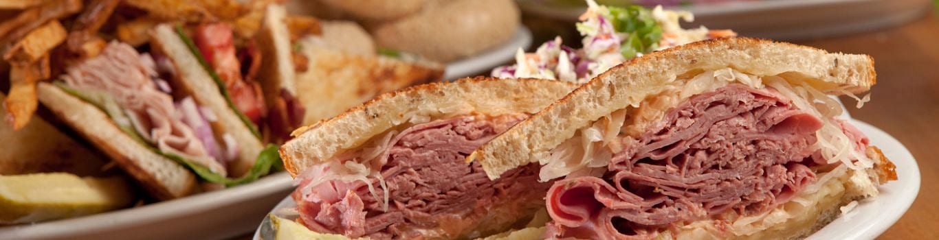 Close-up of a corned beef sandwich with sauerkraut, a club sandwich and fries behind
