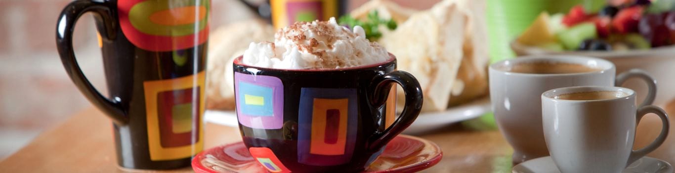 A colourful mug of hot chocolate topped with whipped cream beside coffee cups and pastries